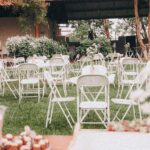 An outdoor wedding venue setup with white folding chairs on green grass and decorative plants.