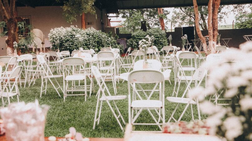 An outdoor wedding venue setup with white folding chairs on green grass and decorative plants.
