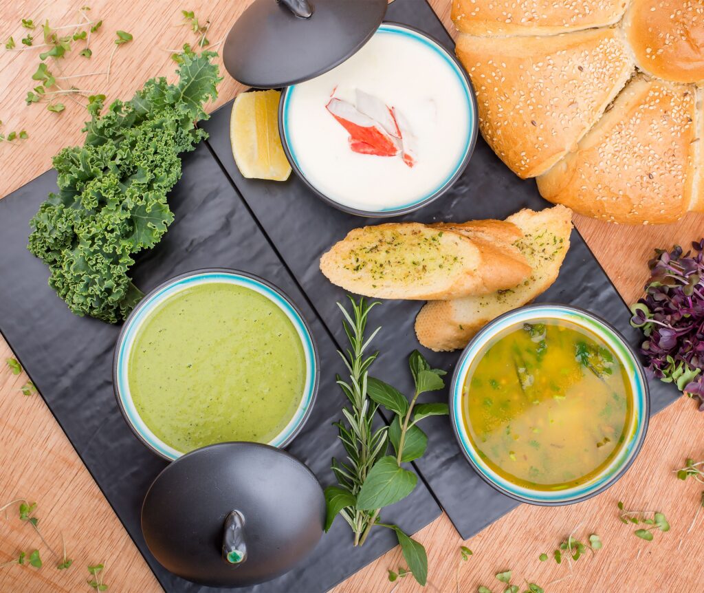 A selection of gourmet soups with fresh bread, herbs, and greens presented artfully on a wooden table.