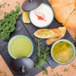 A selection of gourmet soups with fresh bread, herbs, and greens presented artfully on a wooden table.