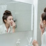 Caucasian woman in bathroom applying skincare product while looking in the mirror.