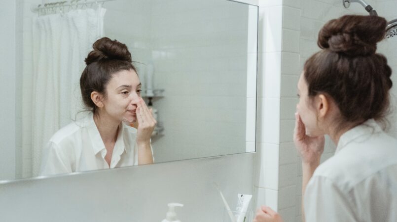 Caucasian woman in bathroom applying skincare product while looking in the mirror.